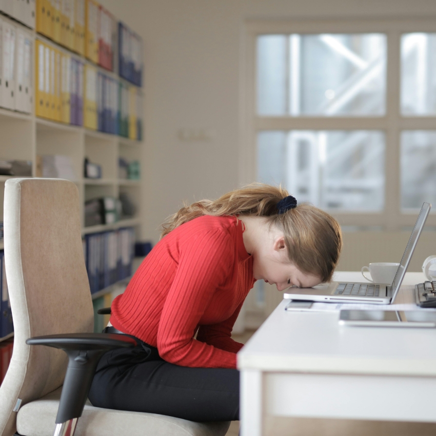 Person in a red sweater rests head on a laptop on a white desk, indicating fatigue or frustration.