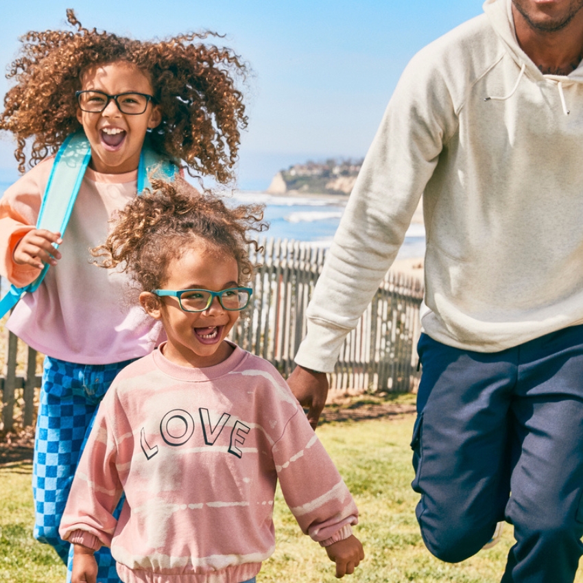 Two children wearing glasses, one in a sweatshirt with "LOVE" text, running and smiling outdoors.