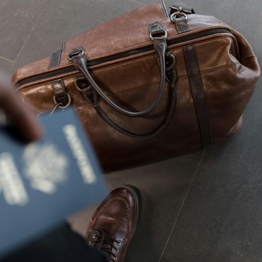 Brown leather duffel bag with double handles and zipper, person holding a blurred passport and wearing brown shoes.