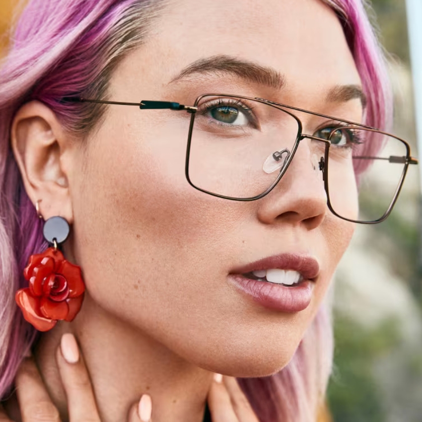 Woman wearing oversized, square-shaped eyeglasses with thin black frames and large, red rose earrings.