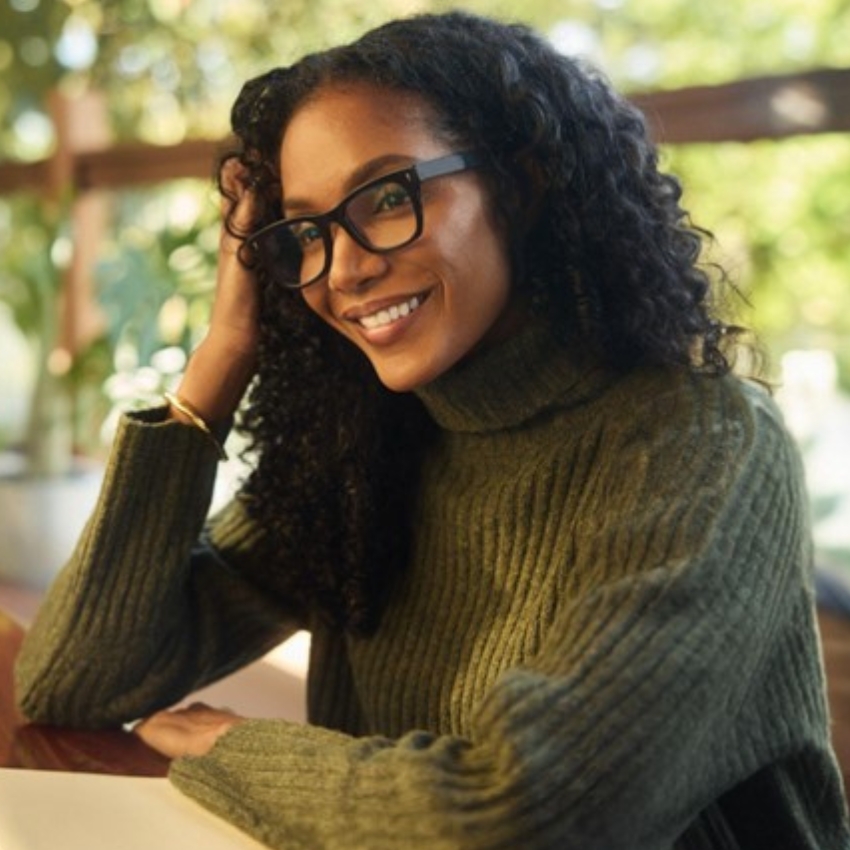 Woman wearing black glasses and a green ribbed turtleneck sweater, smiling and leaning on her hand.