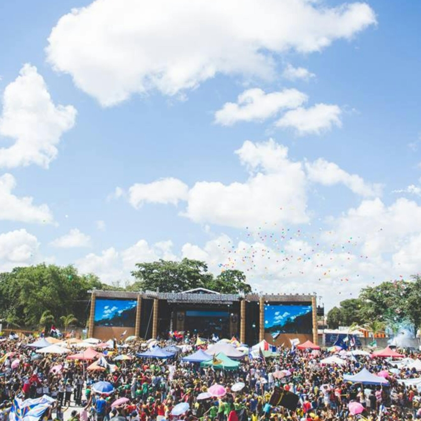 Outdoor festival with large crowd, colorful tents, stage with screens, clear sky filled with scattered clouds.