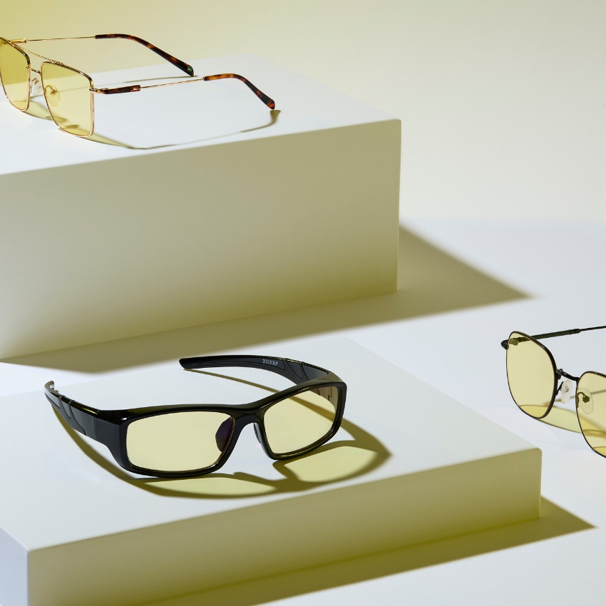 Four pairs of various styles of yellow-tinted glasses are displayed on white platforms.