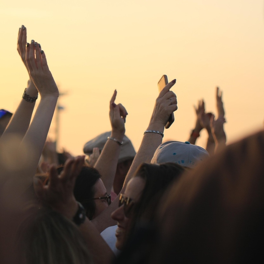 Crowd of people with raised hands, one holding a smartphone, at an outdoor event during sunset.