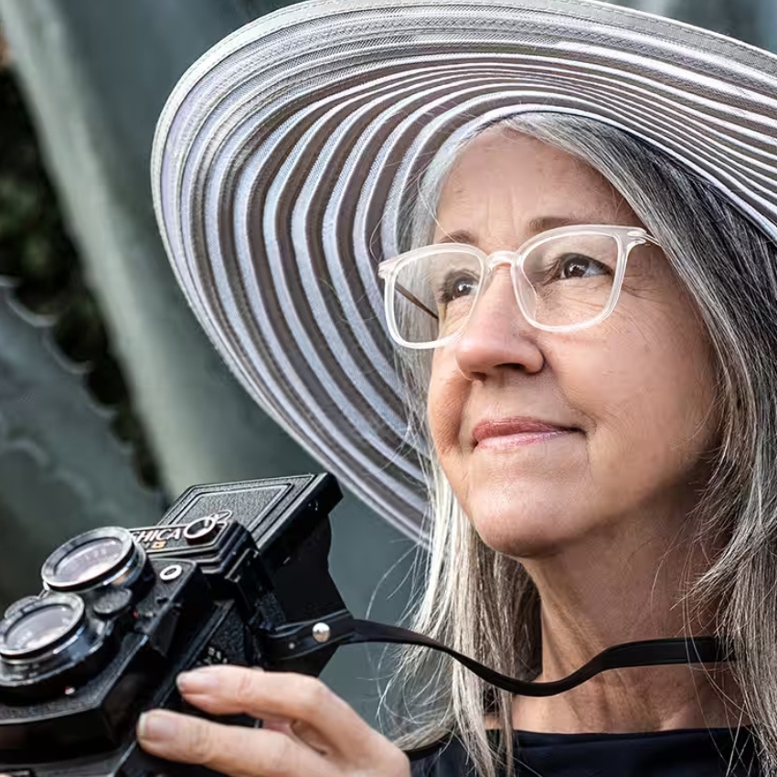 Woman with glasses wearing a wide-brimmed, striped sunhat, holding a vintage Yashica camera.