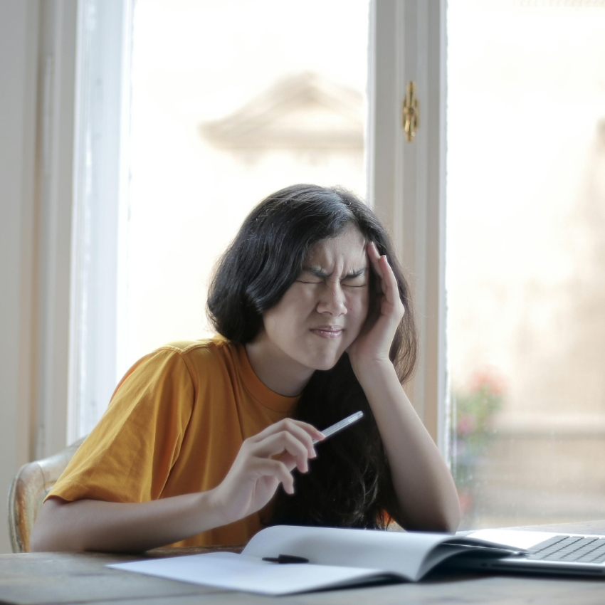 Person in a yellow shirt holding a pen, appearing to have a headache while sitting in front of a laptop.