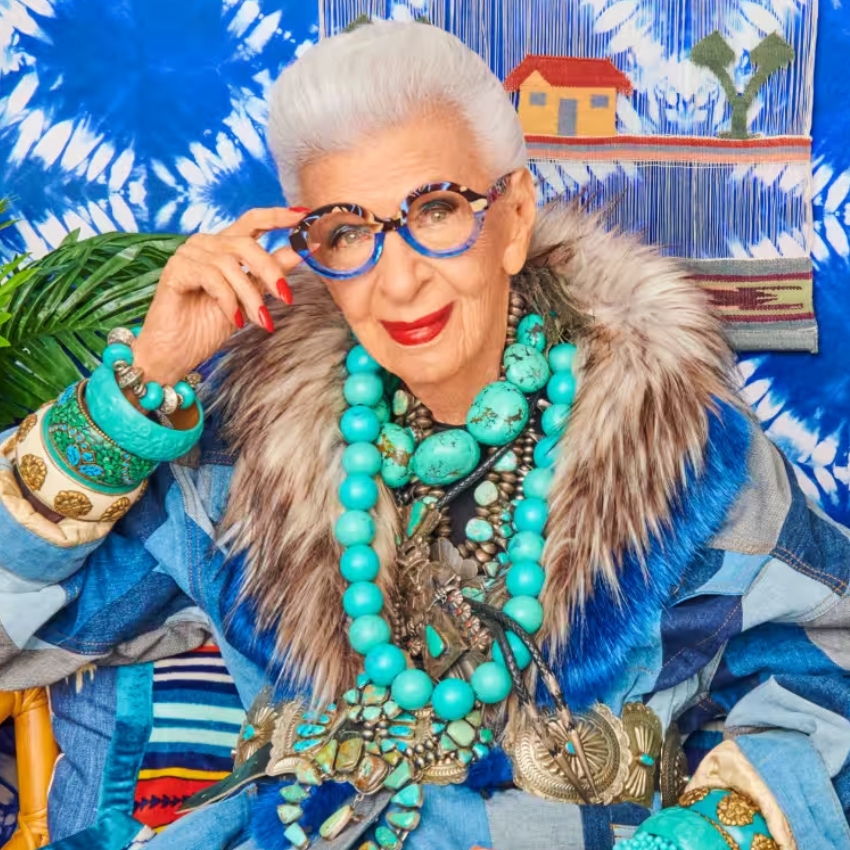 Elderly woman wears large blue beaded necklace, matching glasses, colorful bangles, and a fur-collared coat.