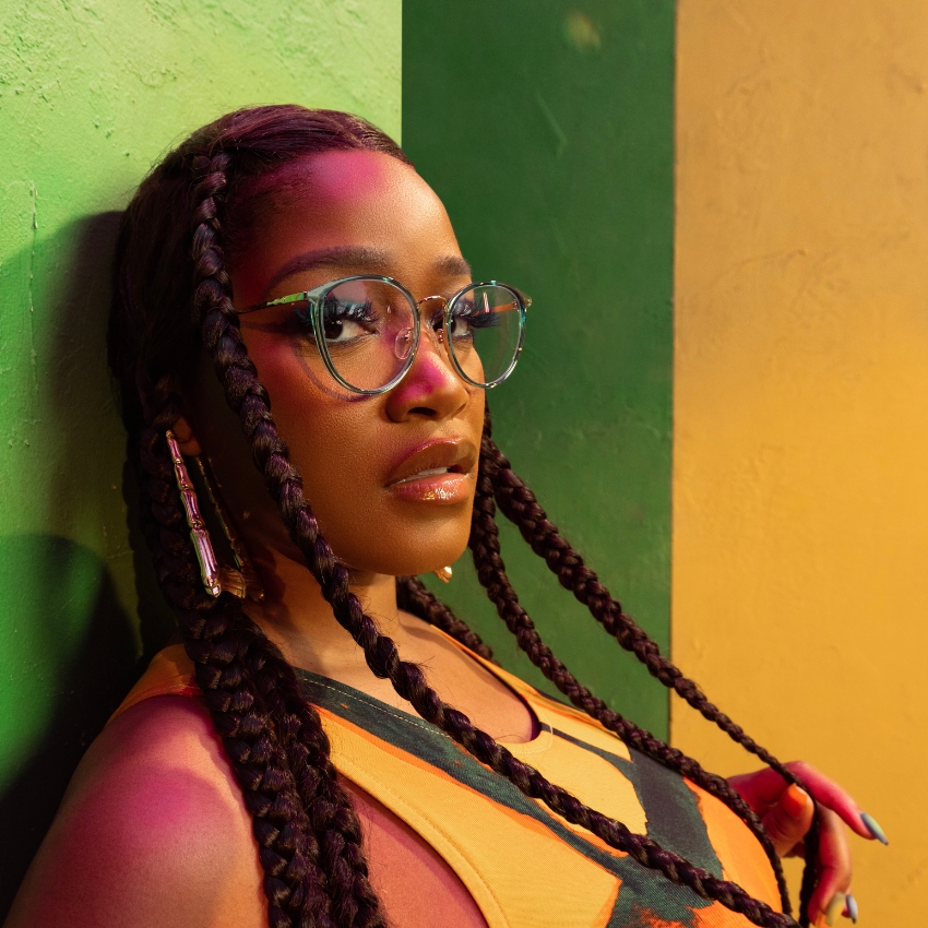 Woman wearing large round eyeglasses with clear frames, long braided hair, and orange top.