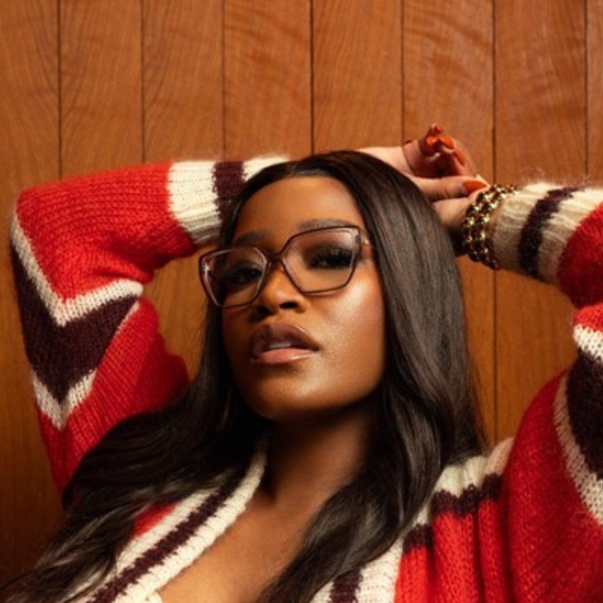 Woman wearing oversized glasses and a red, brown, and white striped sweater against a wooden-paneled background.