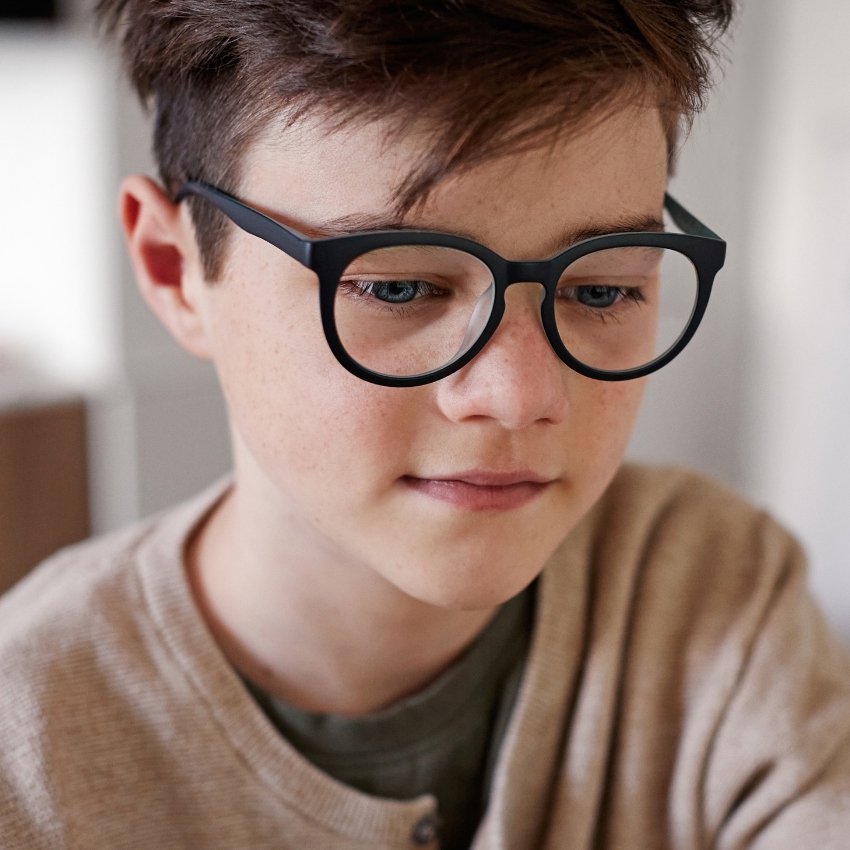 Young boy wearing black round eyeglasses and a beige sweater.