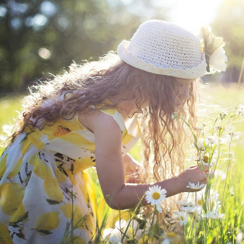 Child wearing a yellow floral dress and a white straw hat with a flower, picking daisies in a field.