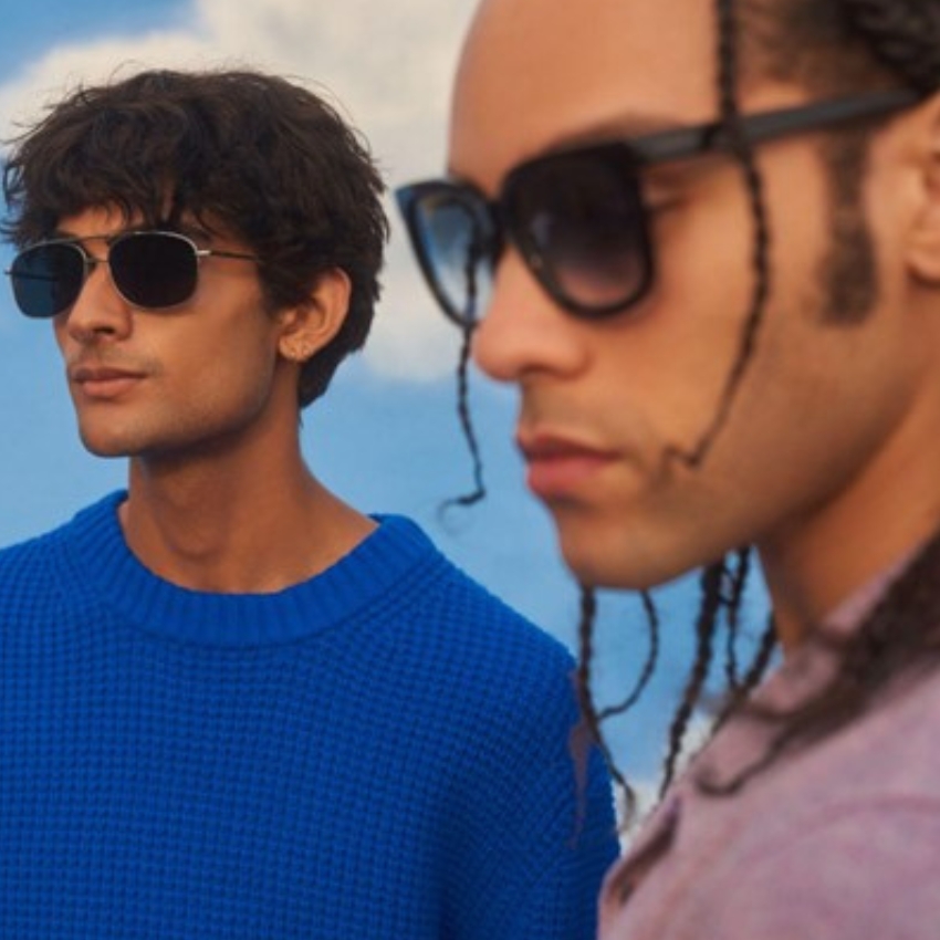 Two men wear stylish black sunglasses, one in a blue sweater and the other in a mauve shirt.