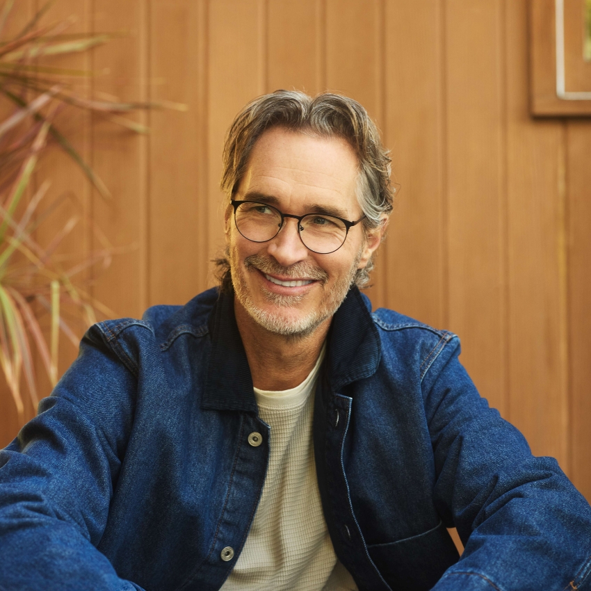 Man wearing glasses, denim jacket, and white shirt, smiling while sitting against a wooden wall background.