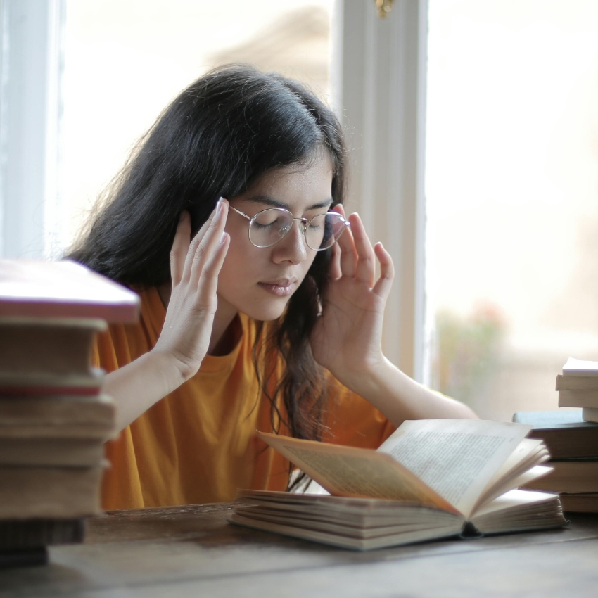 Person reading a book, wearing glasses and a yellow shirt, with hands touching the temples of their head.