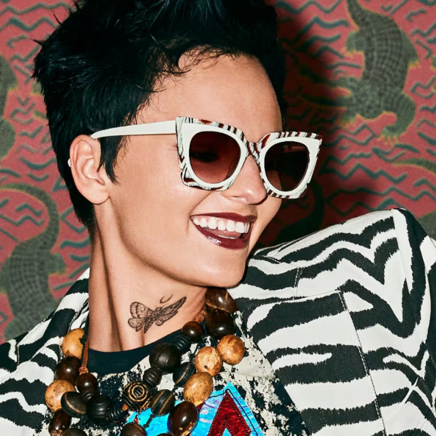 Woman wearing zebra-striped sunglasses and matching jacket, with a butterfly tattoo on neck and beaded necklace.