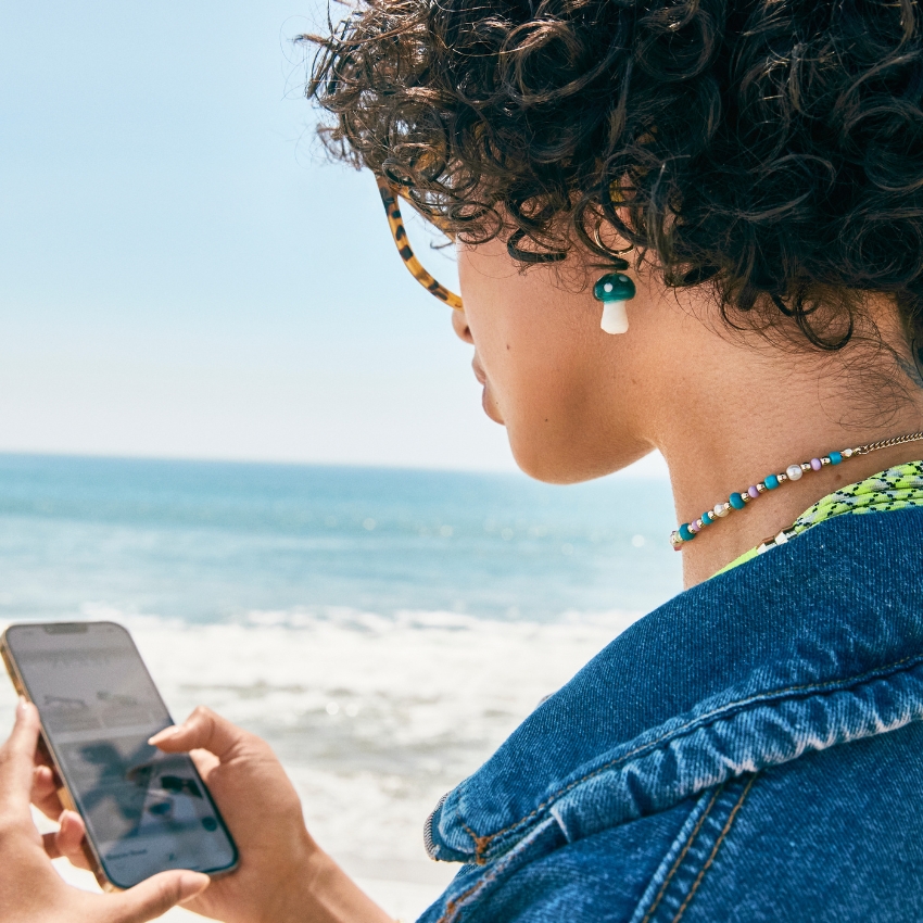 Person wearing turquoise and white earrings, beaded necklace, and holding a smartphone by the ocean.