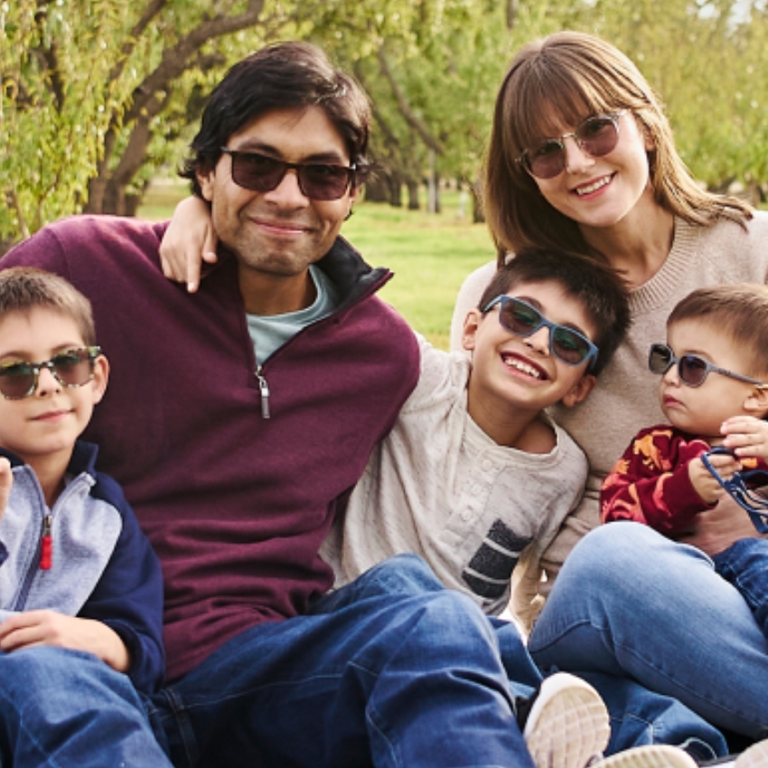 Family of five wearing sunglasses, sitting closely together outdoors.