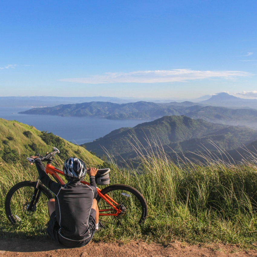 Person with an orange mountain bike sits at a grassy viewpoint, overlooking a scenic mountain landscape.