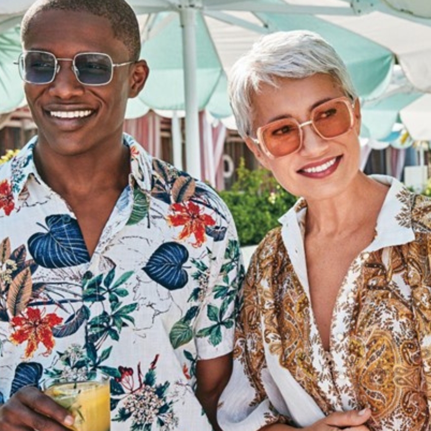 Two people wearing vibrant, patterned shirts and sunglasses, smiling outdoors. One holds a yellow drink.