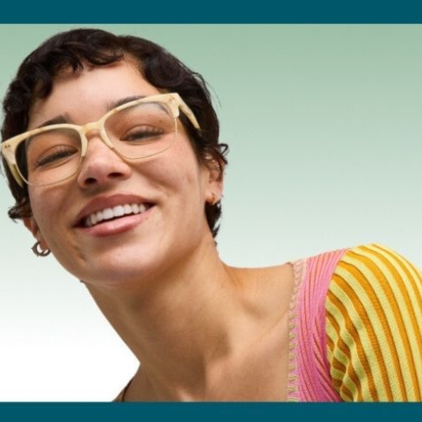 Woman smiling, wearing cream-colored eyeglasses and a striped yellow and pink top against a green background.