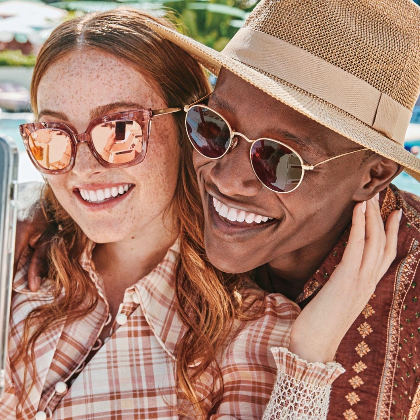 Two people wearing stylish sunglasses, one in pink reflective lenses and the other in round gold-rimmed lenses.