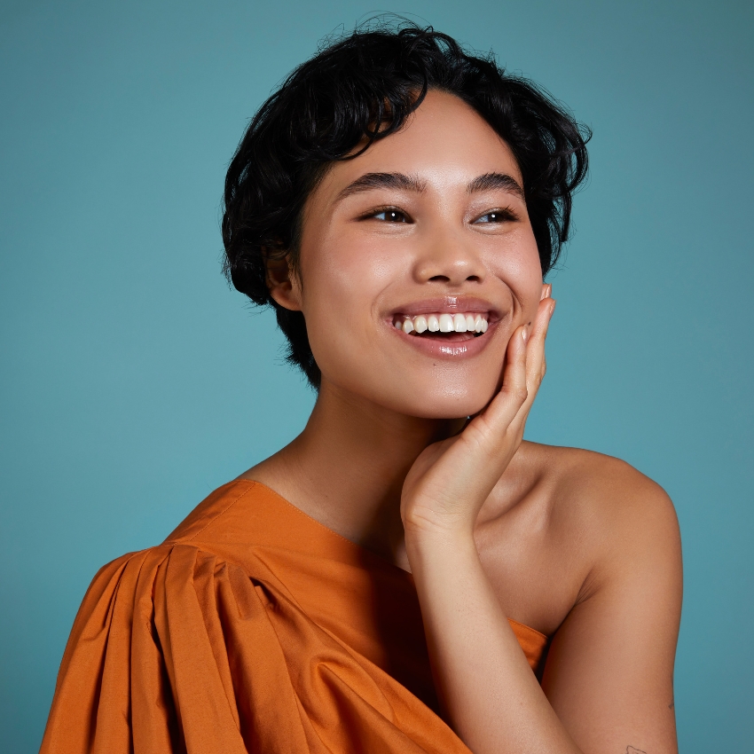 Person in an orange dress with a happy expression and hand on cheek.