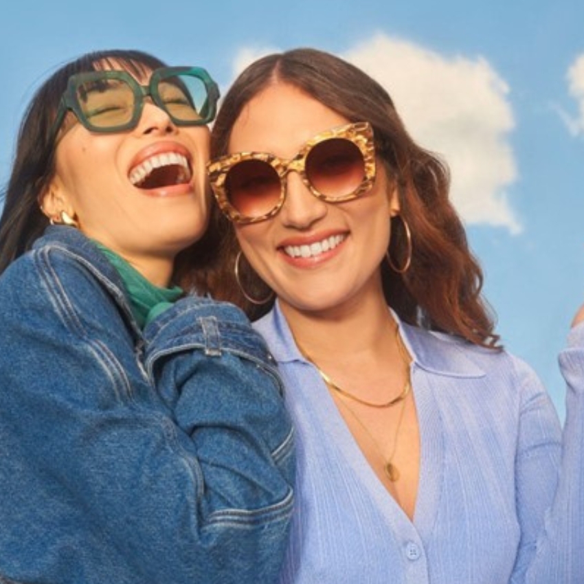 Two women smiling and wearing fashionable sunglasses; one in green frames, the other in tortoiseshell frames.