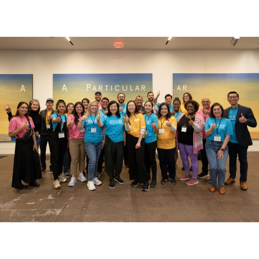 Group of people smiling and giving thumbs-up, wearing colorful shirts with name tags in front that partially reads "A PARTICULAR".