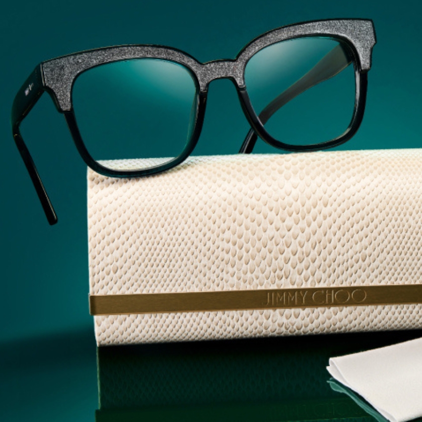 Black square-framed glasses with glitter accents on a beige textured Jimmy Choo case with gold logo band.