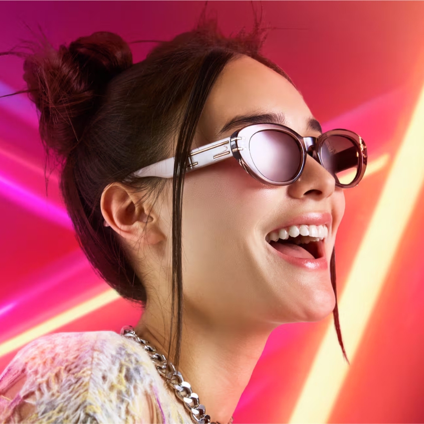 Woman wearing stylish round sunglasses with white frames, smiling against a vibrant neon background.