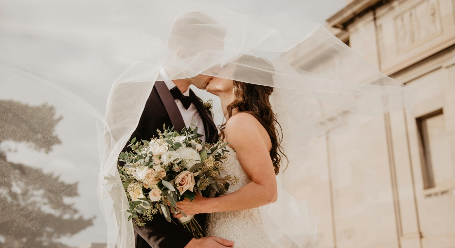 Bride and groom under a flowing veil, bride holding a bouquet of white and pale pink flowers with greenery.