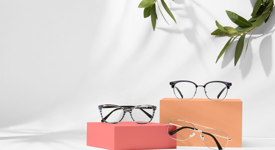 Three pairs of eyeglasses on peach and coral blocks, with leafy green shadows in the background.