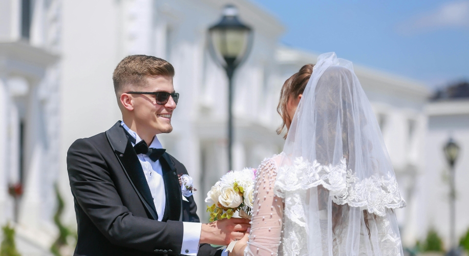 Groom in a black tuxedo and bride in a white lace veil holding a bouquet of white and pink flowers.