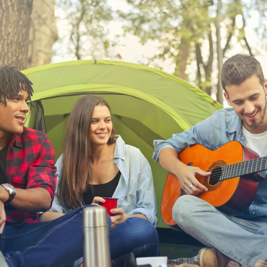 Friends enjoying camping; one plays an orange acoustic guitar, others sit and smile near a green tent.