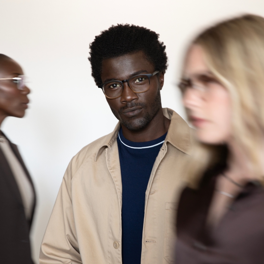 Man wearing dark-rimmed glasses and a beige jacket, with two blurred people in the foreground.