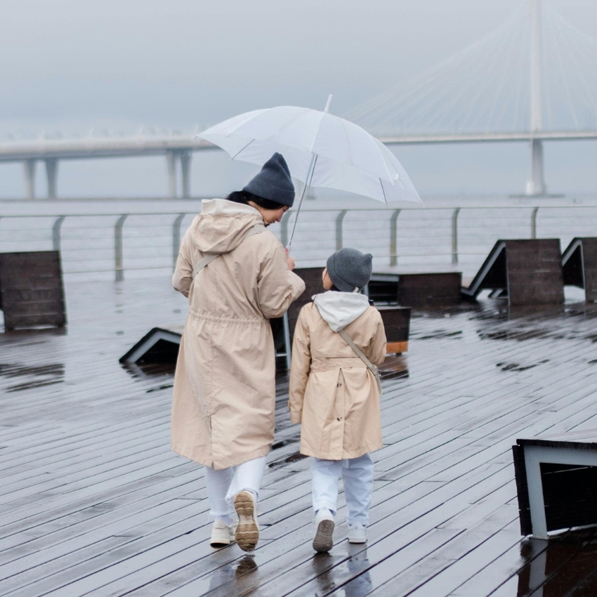 Adult and child wearing beige raincoats and grey knit hats, holding a clear umbrella, walking on a rainy boardwalk.