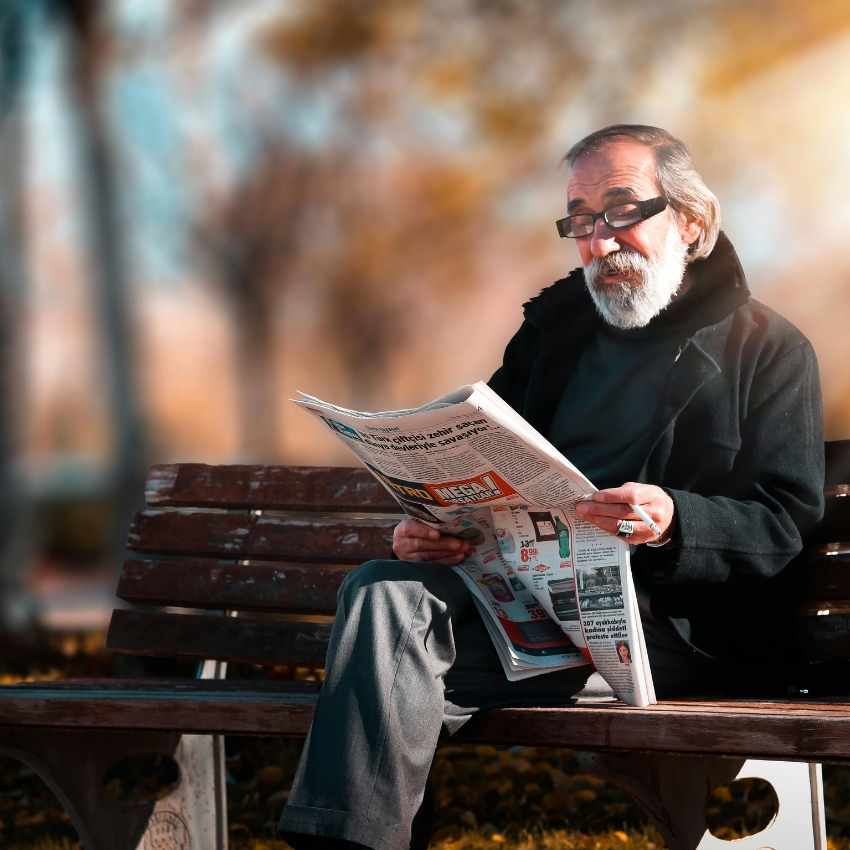 Elderly man in a black coat and glasses reading a newspaper while sitting on a park bench.