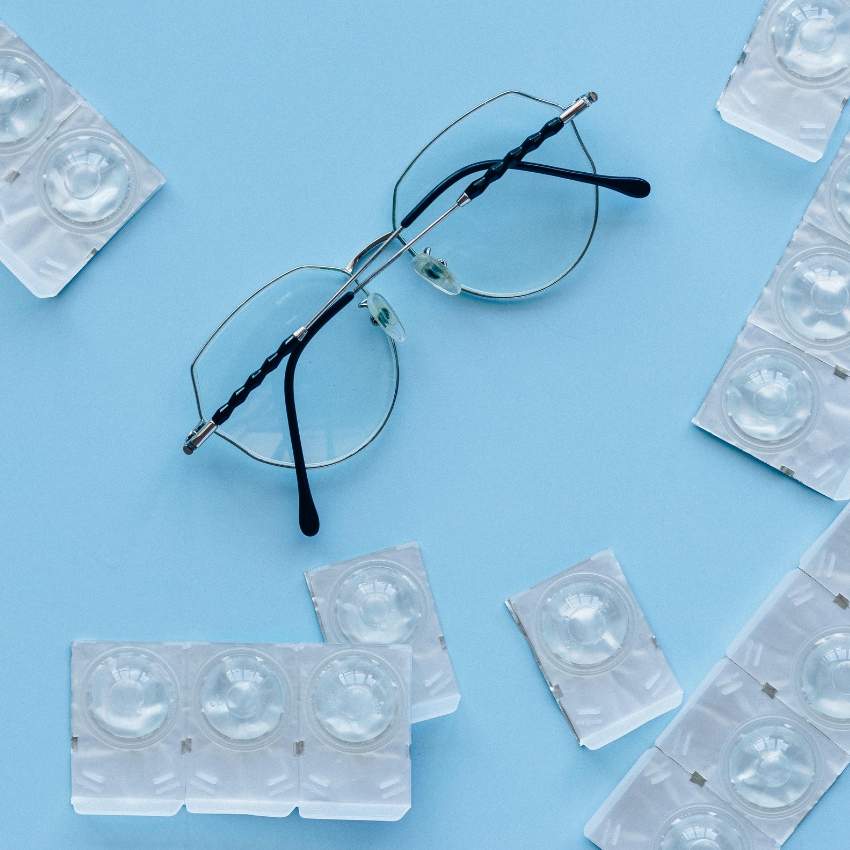 Eyeglasses with various blister packs of contact lenses on a light blue background.