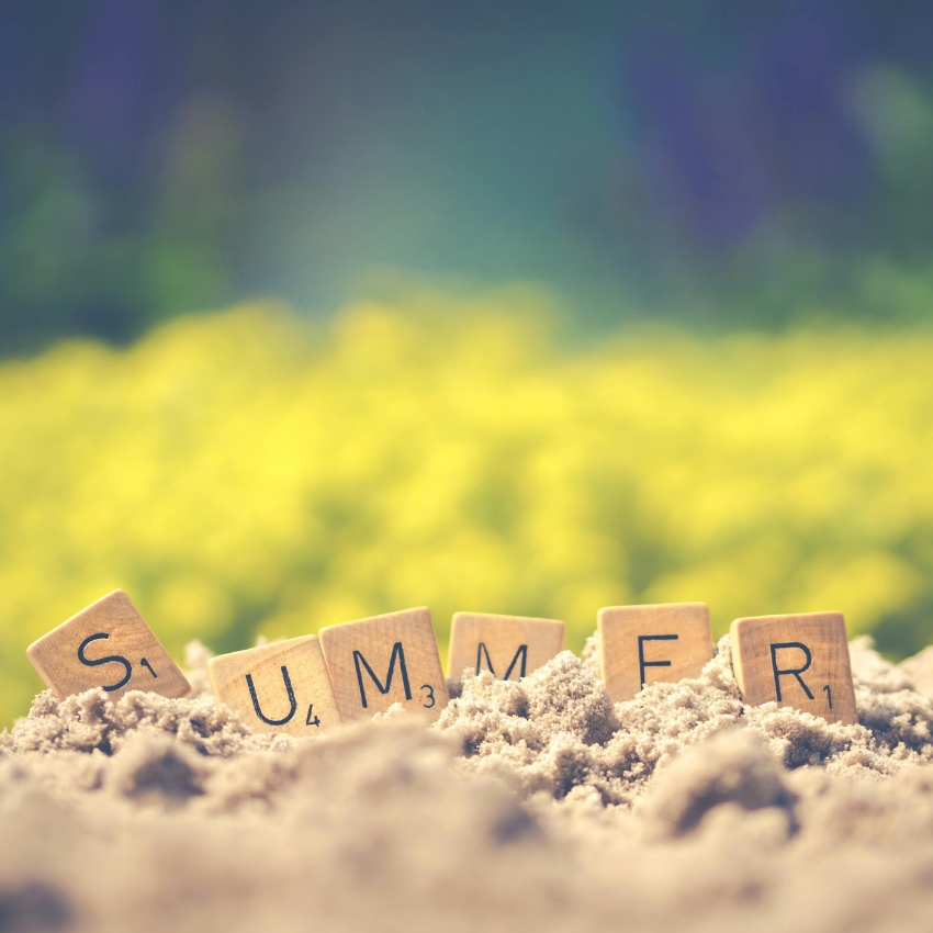 Scrabble tiles spelling "SUMMER" partially buried in sand.