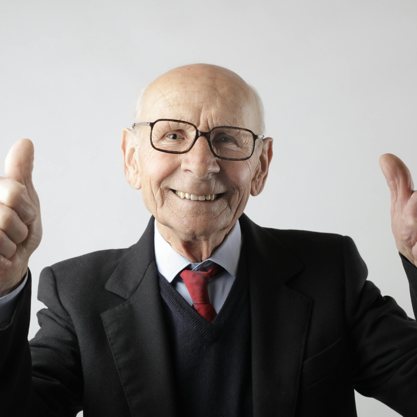 Elderly man in a suit and red tie giving thumbs up while smiling.