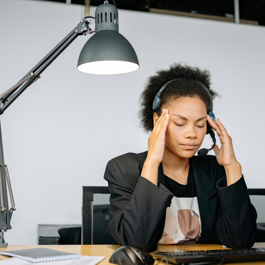 Person wearing a headset, seated at a desk with a computer, and touching temples with a stressed expression.