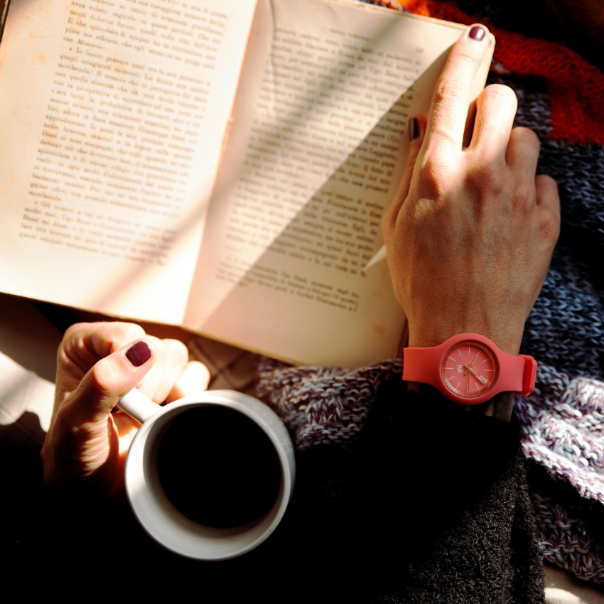 Hand with coral pink watch, finger pointing on open book with other hand holding a cup of coffee.