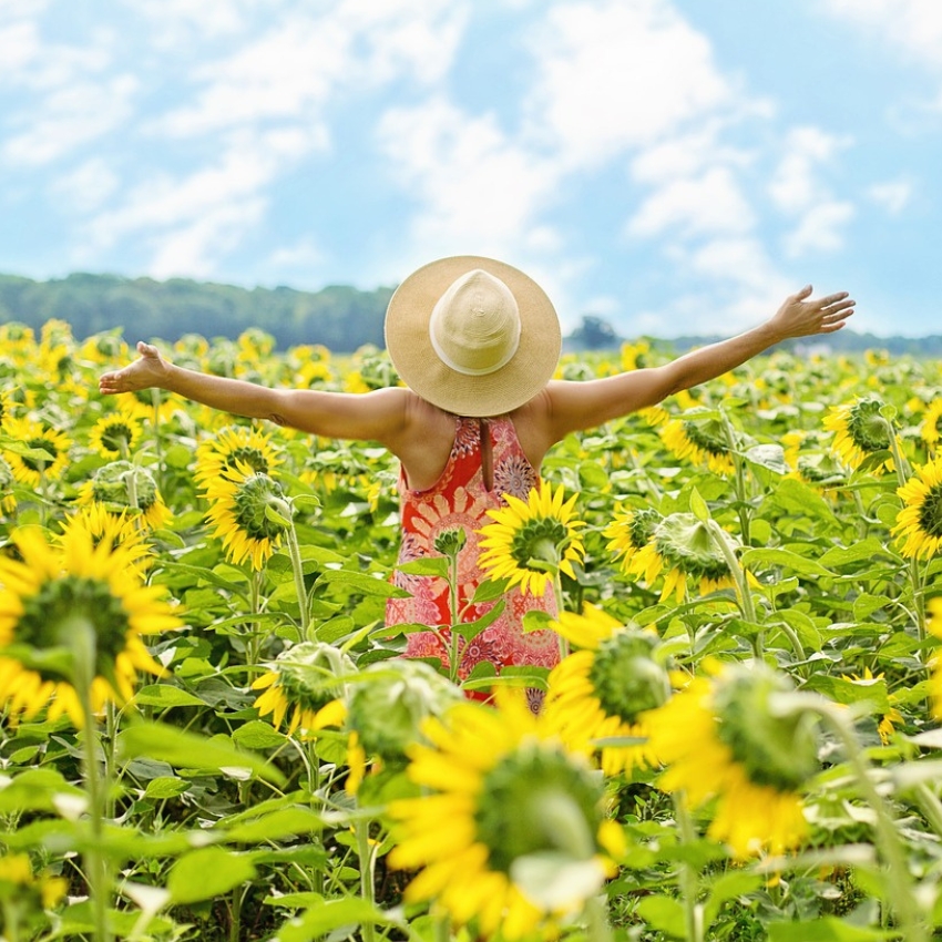 Person in a straw hat and floral dress stands in a sunflower field, arms outstretched to the sky.