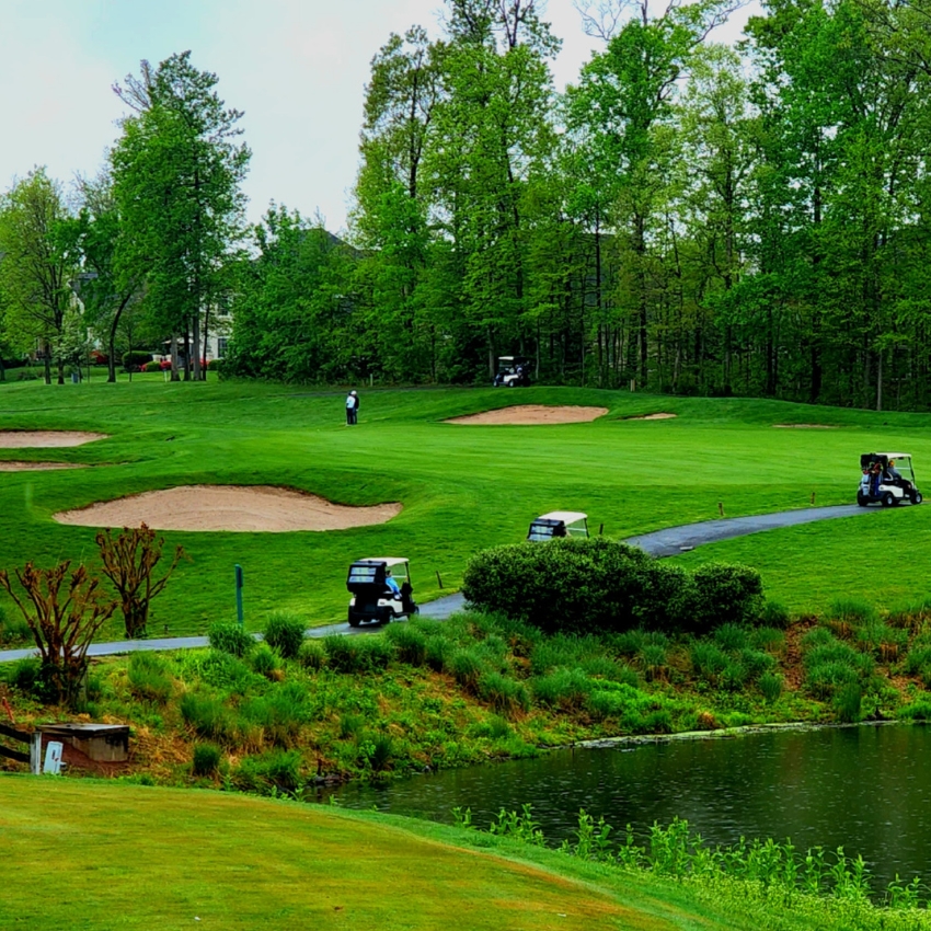 Golf course with sand traps, golfers in the distance, and golf carts on a paved path near a small pond.