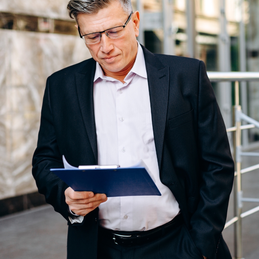 Man in a suit holding a blue clipboard and reading a document outdoors.