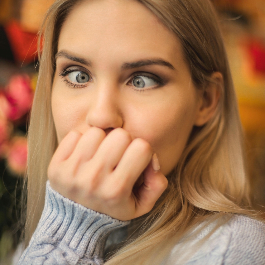 Blonde woman in light blue sweater with wide eyes and hand covering her mouth, appearing surprised or shocked.