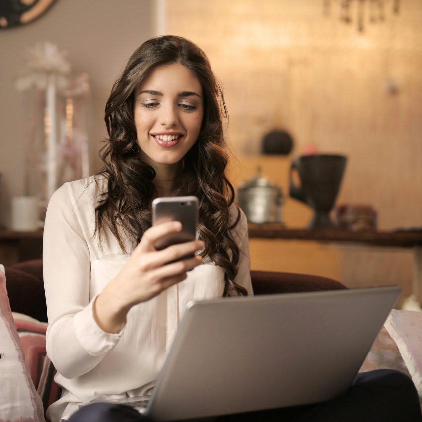 Woman using a smartphone and laptop, seated on a couch.