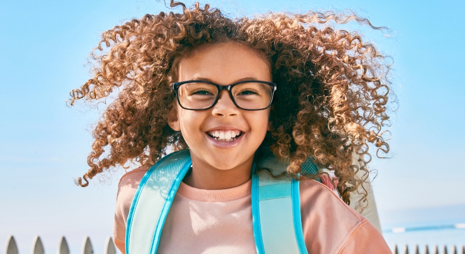 Smiling child with curly hair, wearing black glasses and a turquoise backpack.