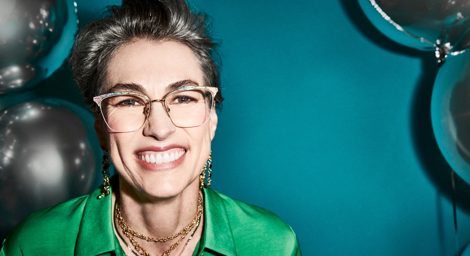 Woman with short gray hair wearing oversized, gold-framed glasses, green shirt, and layered necklaces smiling.