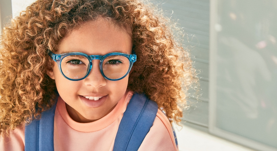 Child with curly hair wearing blue eyeglasses and a pink sweater.